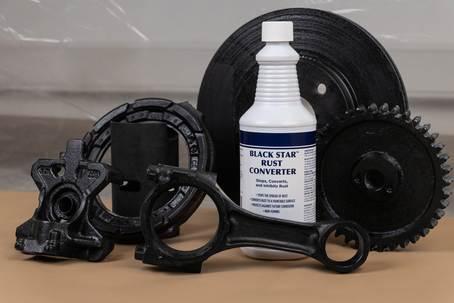 Bottle of Black Star Rust Converter surrounded by various restored black metal parts, including gears, a brake rotor, and automotive components.