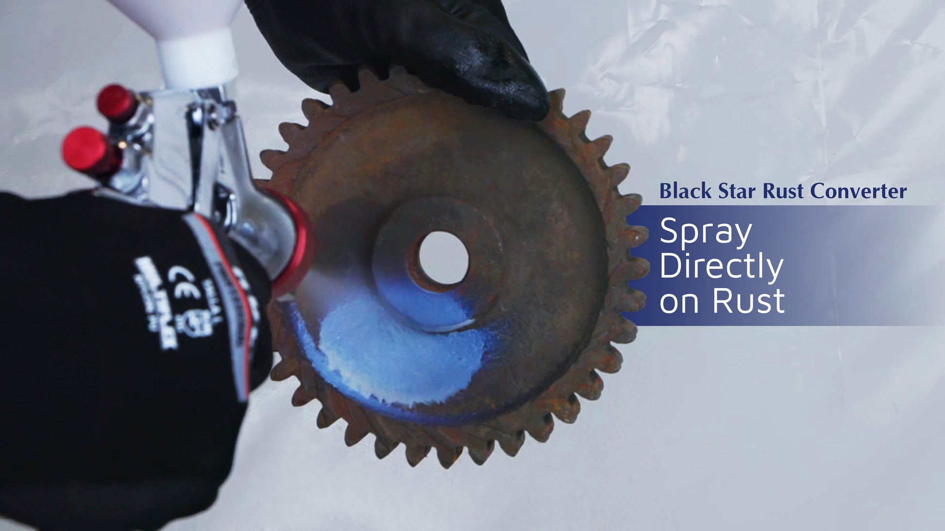 Gloved hands spraying Black Star Rust Converter onto a rusty gear, with the blue spray covering part of the rusted surface