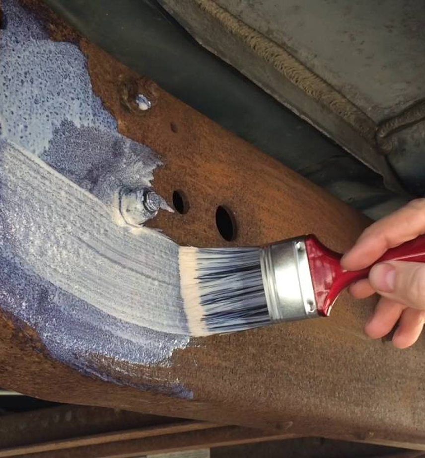 Hand using a paintbrush to apply a blue-tinted rust converter onto a rusted metal surface with bolt holes.