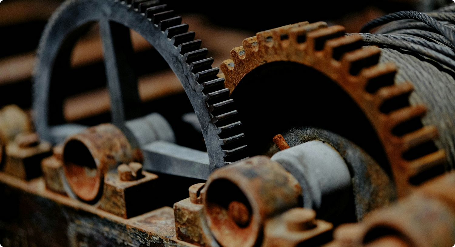 Close-up of interlocking metal gears and pulleys, partially covered in rust, with a steel cable wound around one gear.